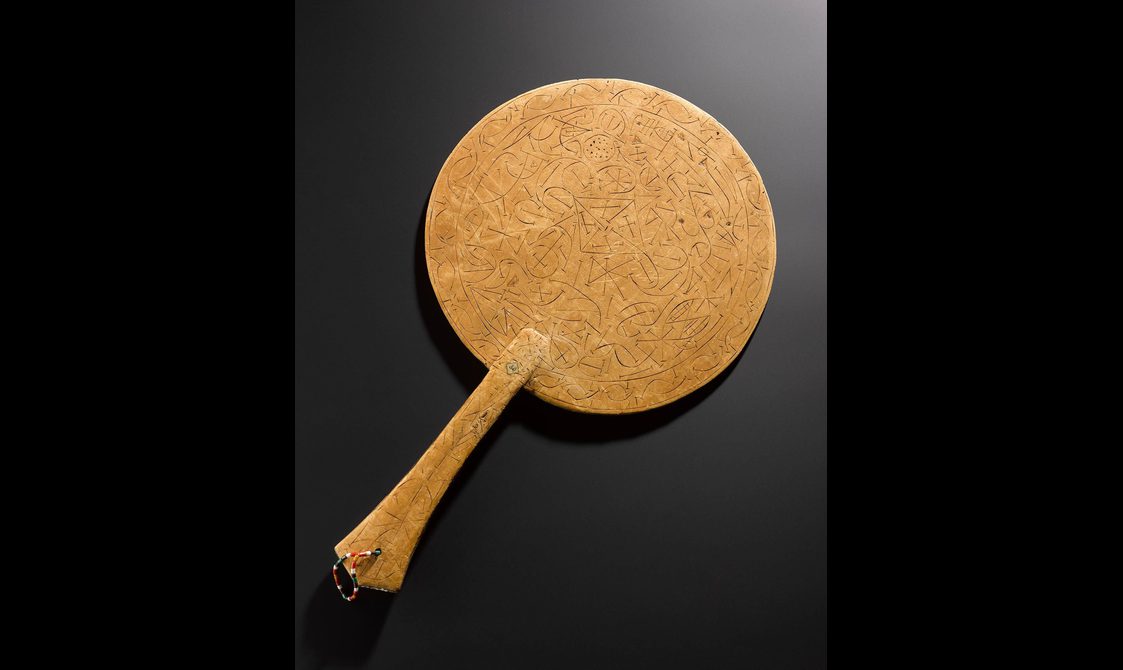 A wooden circular fan with a handle. The fan has cross patterns carved into it.