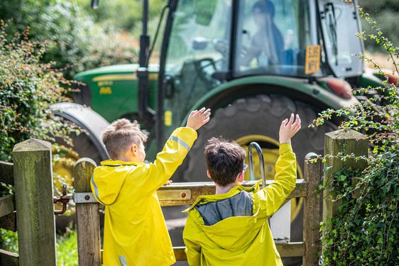 Two children in yellow jackets waving at a green tractor.