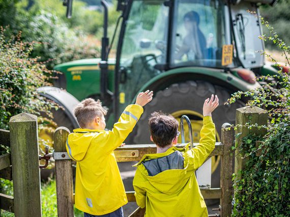Two children in yellow jackets waving at a green tractor.