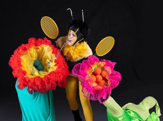 A person dressed as a bee sips through a straw from brightly coloured flowers. The flowers are also people in costume.