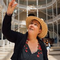 Woman with a large straw hat reaching upwards with her right hand, in a large Victorian gallery.