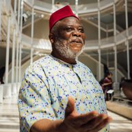 Man wearing traditional Ghanian outfit with his hands outstretched in a large Victorian gallery.