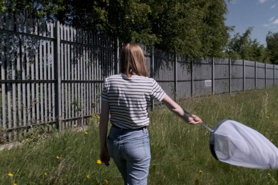 A person with long blonde hair and a white striped shirt seend from behind holds a large insect net over tall grass while walking along a fence.