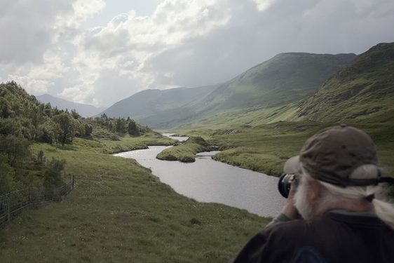 Looking over the shoulder of a person in the foreground photographing a vast green valley with a river running through the centre, a forest on the left, and mountains in the distance.