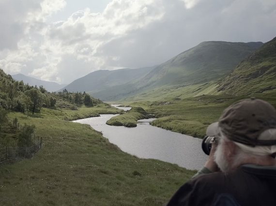 Looking over the shoulder of a person in the foreground photographing a vast green valley with a river running through the centre, a forest on the left, and mountains in the distance.