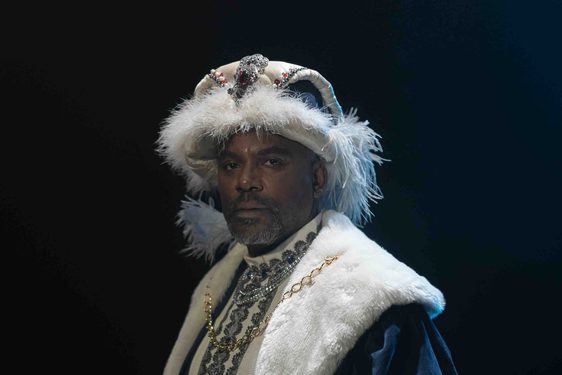 Film still of a 16th century Black African diplomat dressed in elaborate European-style clothing with a thick, white fur trimmed collar and hat. He is holding a scroll.