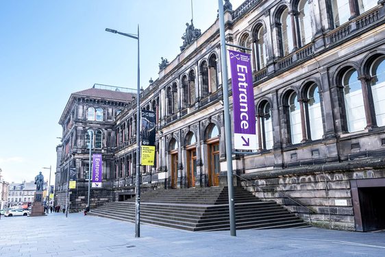 Wide shot of a large brown museum building with grand windows. A statue of a man stands outside the entrance doors.