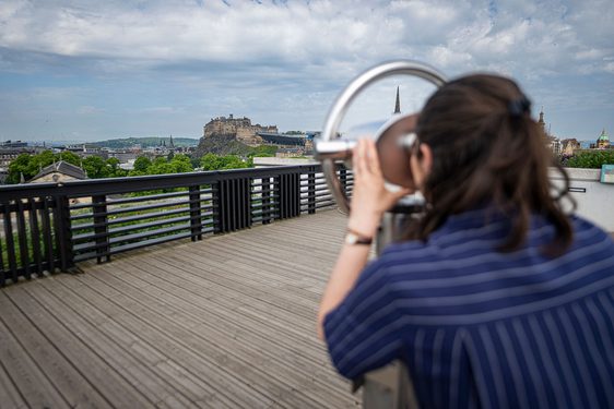 A woman wearing a stripey jumpsuit looks through a view finder out towards the Edinburgh skyline. In view is Edinburgh Castle.