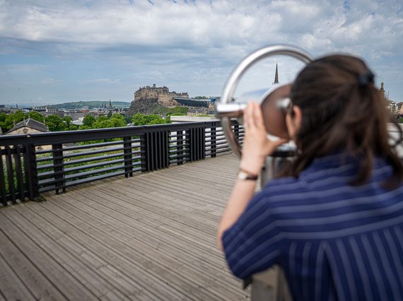 A woman wearing a stripey jumpsuit looks through a view finder out towards the Edinburgh skyline. In view is Edinburgh Castle.