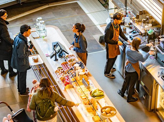 Customers queueing at a cafe till. Baristas are making coffees.