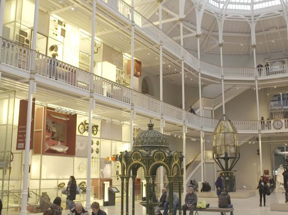 A multi-level museum atrium with white pillars and railings. Different museum objects are on display on each level. Visitors look up at the objects.