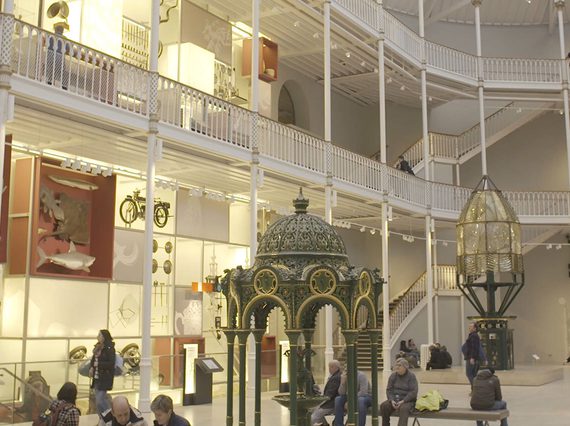 A multi-level museum atrium with white pillars and railings. Different museum objects are on display on each level. Visitors look up at the objects.