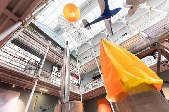 Two orange hot air balloons in a large museum gallery. The balloon on the left is inflated, the one on the right is delfated.