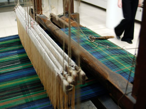 Looking down on a loom mid-weave of a length of blue and green tartan cloth.