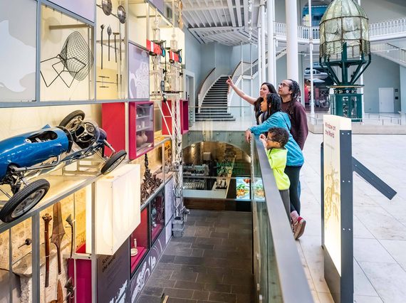 A family looking and pointing at objects mounted in rows on the wall of a museum gallery.