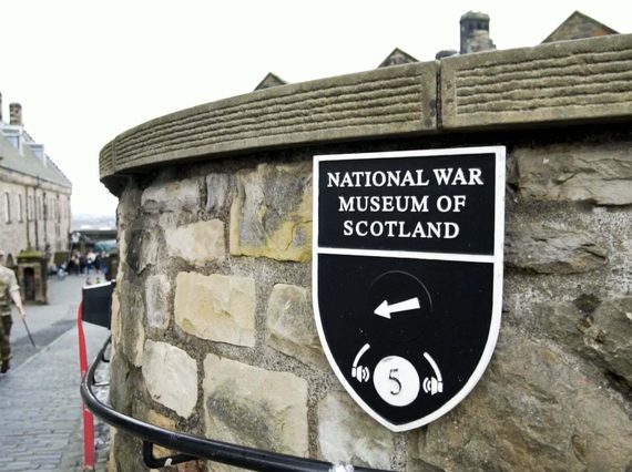 A sign for the National War Museum of Scotland on a brick wall. In the distance a soldier in uniform is walking towards the entrance of the museum.