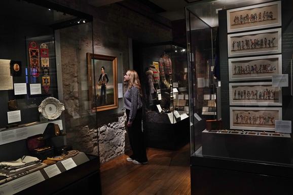 A person with long blonde hair looking at a portrait of a Scottish soldier, surrounded by several glass museum cases.