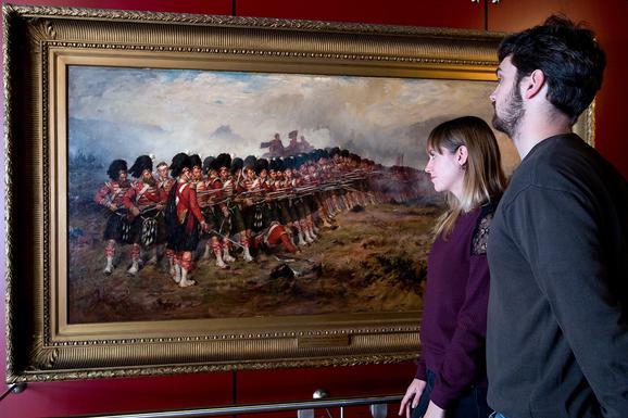 A couple stand looking at a landscape portrait of Scottish soldiers in traditional army uniform. The soldiers are standing in a line waiting to charge in battle.