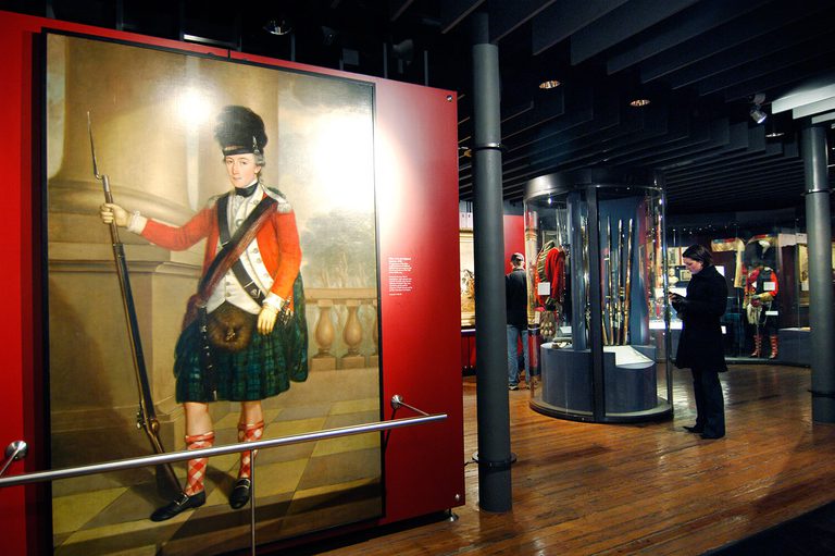 A museum gallery with a large portrait of a Scottish soldier in traditional uniform dress. There are visitors in the background looking at objects in cases.