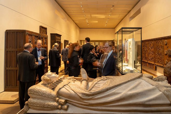 A group of people at a drinks reception in a museum gallery. At the front is a replica of the casket of Mary Queen of Scots. Lining the walls are ornate pieces of wooden furniture.