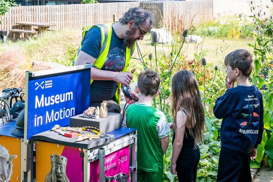 A member of the museum learning team shows three children some museum handling objects. They are standing next to a cargo bike with the words ‘Museum in Motion’ on it.
