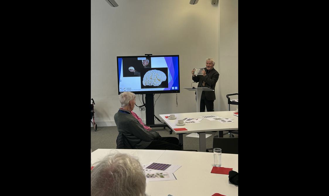 A man stands in front of a group of people giving a presentation. Next to him is an image of a drawing of a brain.
