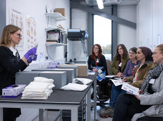 A curator holding a collection item in a museum store. They are giving a talk to a group of visitors who are sitting down.
