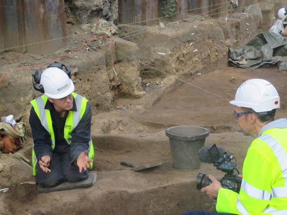 Two archeaologists in high visibility gear in a dig site. One is kneeling down and talking to a video camera, which the other is holding.