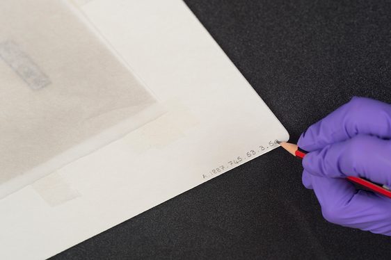 A gloved hand applying a label to a mounted artwork with a pencil.