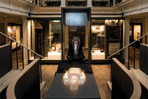 An actor dressed in Victorian clothes standing over a virtual cadaver on a dissection table in an anatomical theatre.