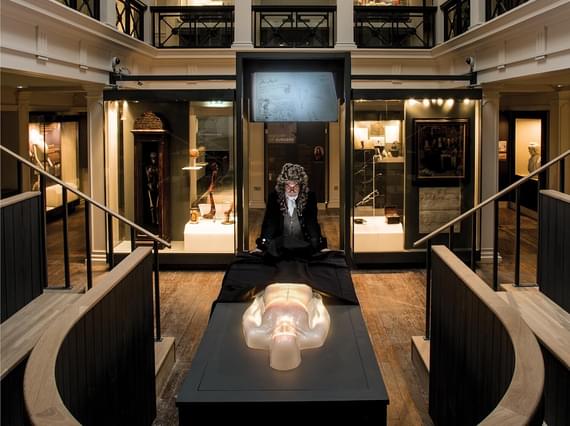 An actor dressed in Victorian clothes standing over a virtual cadaver on a dissection table in an anatomical theatre.