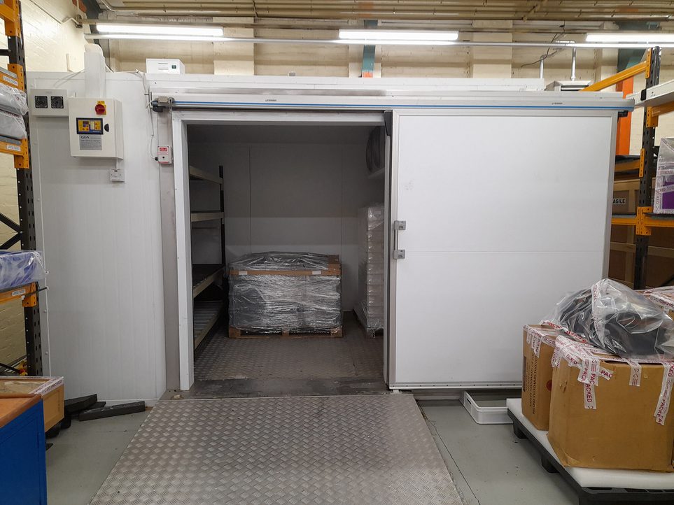 A large walk-in freezer in a museum collection centre. The door of the freezer is open, with a row of shelves inside and a large crate wrapped in plastic wrap.