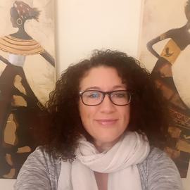 Woman with dark brown hair, grey jumper and white scarf looks forward into the camera with art of two dancing figures behind her.