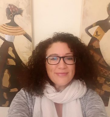 Woman with dark brown hair, grey jumper and white scarf looks forward into the camera with art of two dancing figures behind her.
