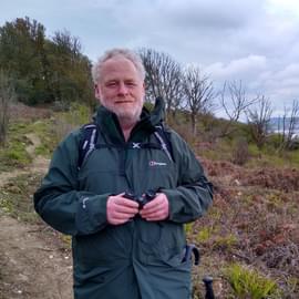 Middle-aged palaeontologist with beard and waterproofs on grey autumn day, standing with binoculars. On unstable landslipped terrain with dead trees, just west of Lyme Regis, Dorset, with the harbour, the sea, and the Dorset sea-cliffs in the distance.