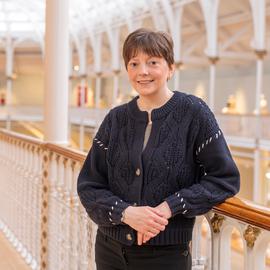 Angela Robb, a white cis woman in her early 40s, with short brown hair. Standing in front of railings in a light-filled museum atrium.
