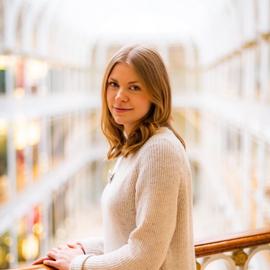 Ashleigh Whiffin, a white woman in her early 30s with dark blonde wavey hair. She is wearing a cream-coloured jumper and standing in front of railings in the grand gallery of the museum.