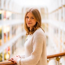 Ashleigh Whiffin, a white woman in her early 30s with dark blonde wavey hair. She is wearing a cream-coloured jumper and standing in front of railings in the grand gallery of the museum.