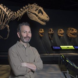 A bearded man standing smiling in front of a dinosaur skeleton