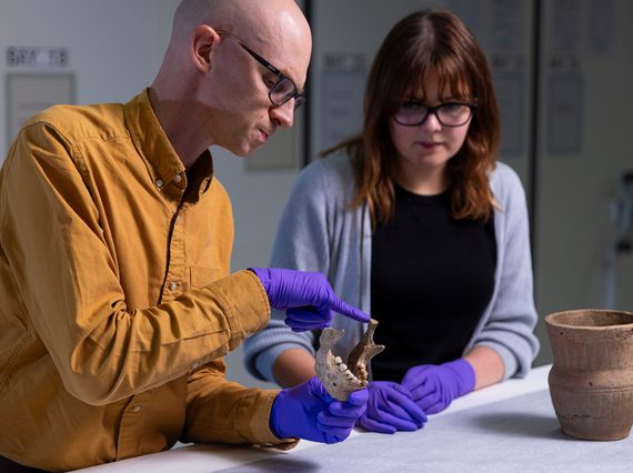2 researchers are looking at a jawbone in a museum storage facility. They're wearing purple gloves, and pointing at features on it.