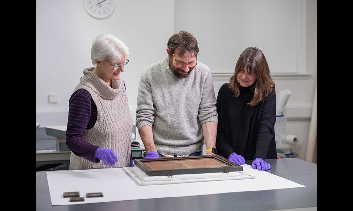 Three people wearing purple gloves standing in a lab, discussing the painting in front of them.