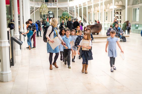 A teacher leads a group of young school children through a large, bright, open gallery.