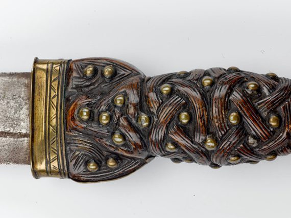 Close-up of an inicately carved dark brown wooden hilt of a dagger, with a bronze coloured metal band at the top edge.