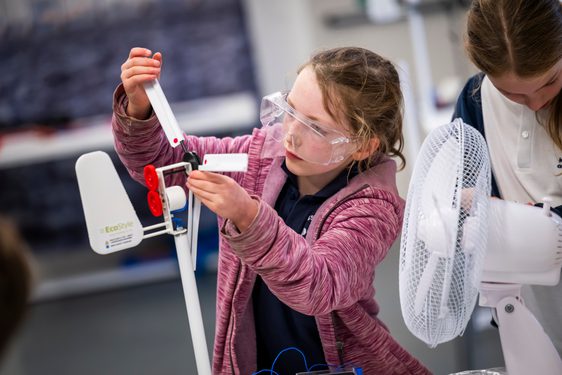 A child in a pink hoodie with long hair tied back wears protective goggles and adjusts a small wind turbine. There is a small fan pointed at the turbine.