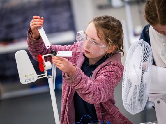A child in a pink hoodie with long hair tied back wears protective goggles and adjusts a small wind turbine. There is a small fan pointed at the turbine.