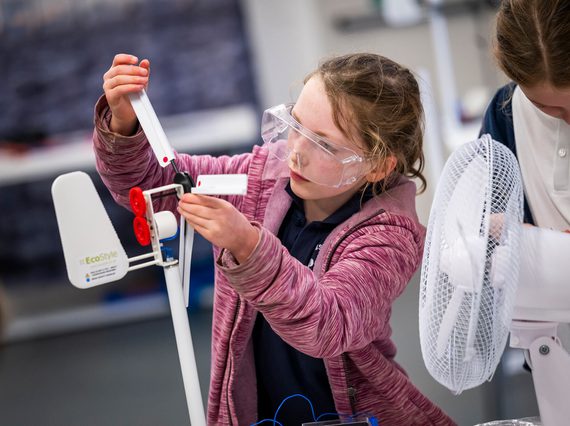 A child in a pink hoodie with long hair tied back wears protective goggles and adjusts a small wind turbine. There is a small fan pointed at the turbine.