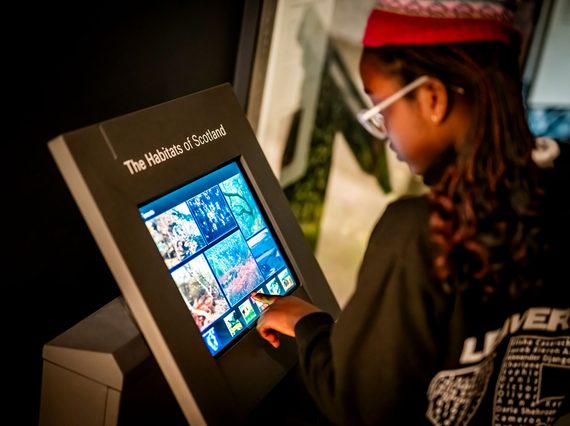 A child with braids and glasses looks at a touchscreen with the title 'The Habitats of Scotland'.