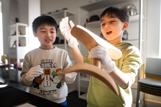 Two children with short dark hair wearing white gloves stand looking at a curved wooden artefact with looks of interest. One of the children is holding the artefact.