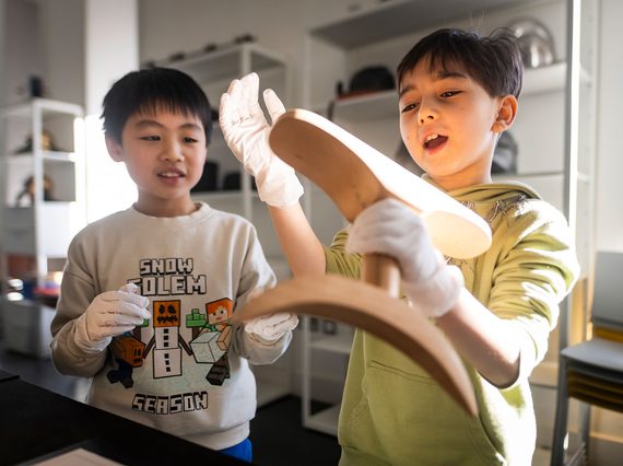 Two children with short dark hair wearing white gloves stand looking at a curved wooden artefact with looks of interest. One of the children is holding the artefact.