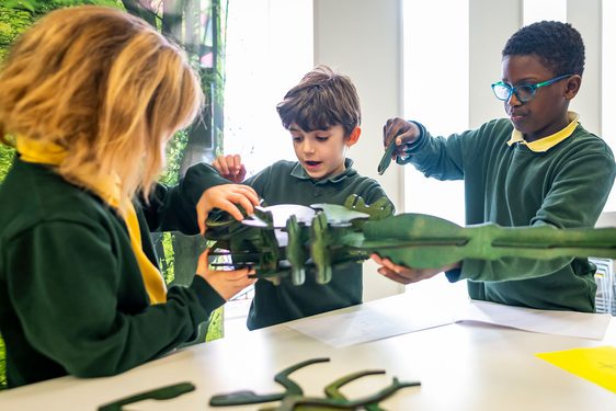 Three children in green tops stand around a table building a model minibeast out of flat, green, wooden parts.